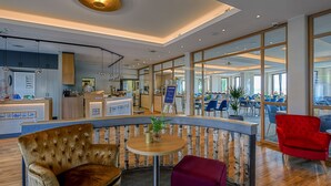 A modern hotel lobby with a curved reception desk, seating area with plush chairs, and large windows offering views of the interior and outside. - BE BIO Hotel be natural (Toenning)