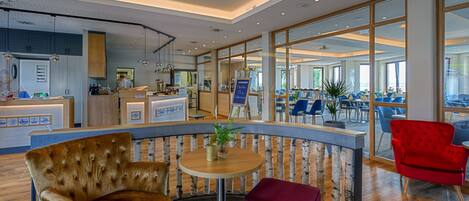 Eine moderne Hotelhalle mit einer gebogenen Rezeption, einer Sitzecke mit bequemen Sesseln und großen Fenstern, die Blicke ins Innere und nach außen ermöglichen.