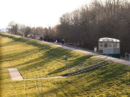 Un parque con una pendiente cubierta de césped, un sendero para caminar, bancos y un pequeño refugio.