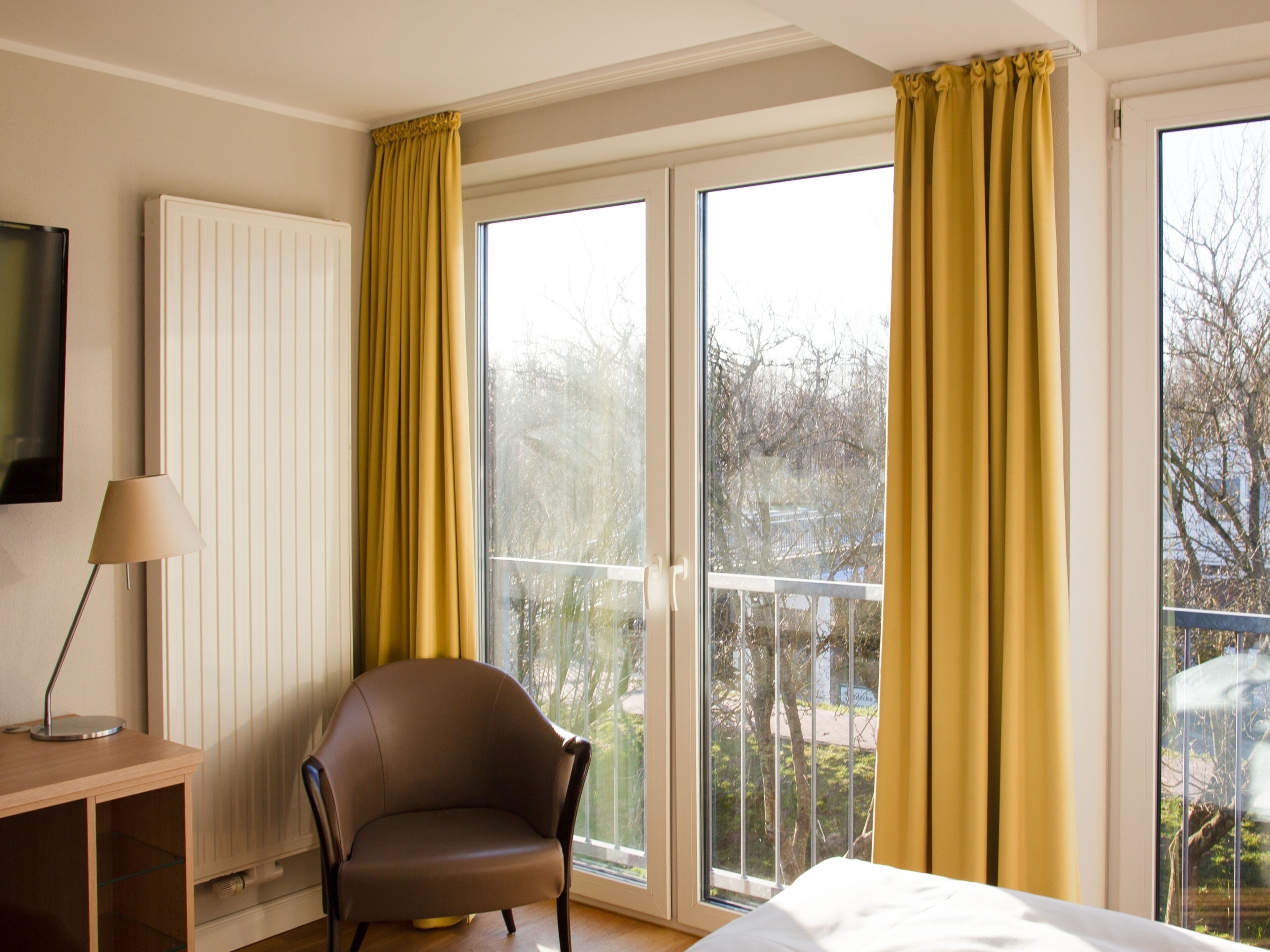 Una habitación con cama, una silla, un escritorio, una lámpara y ventanas amplias con cortinas.