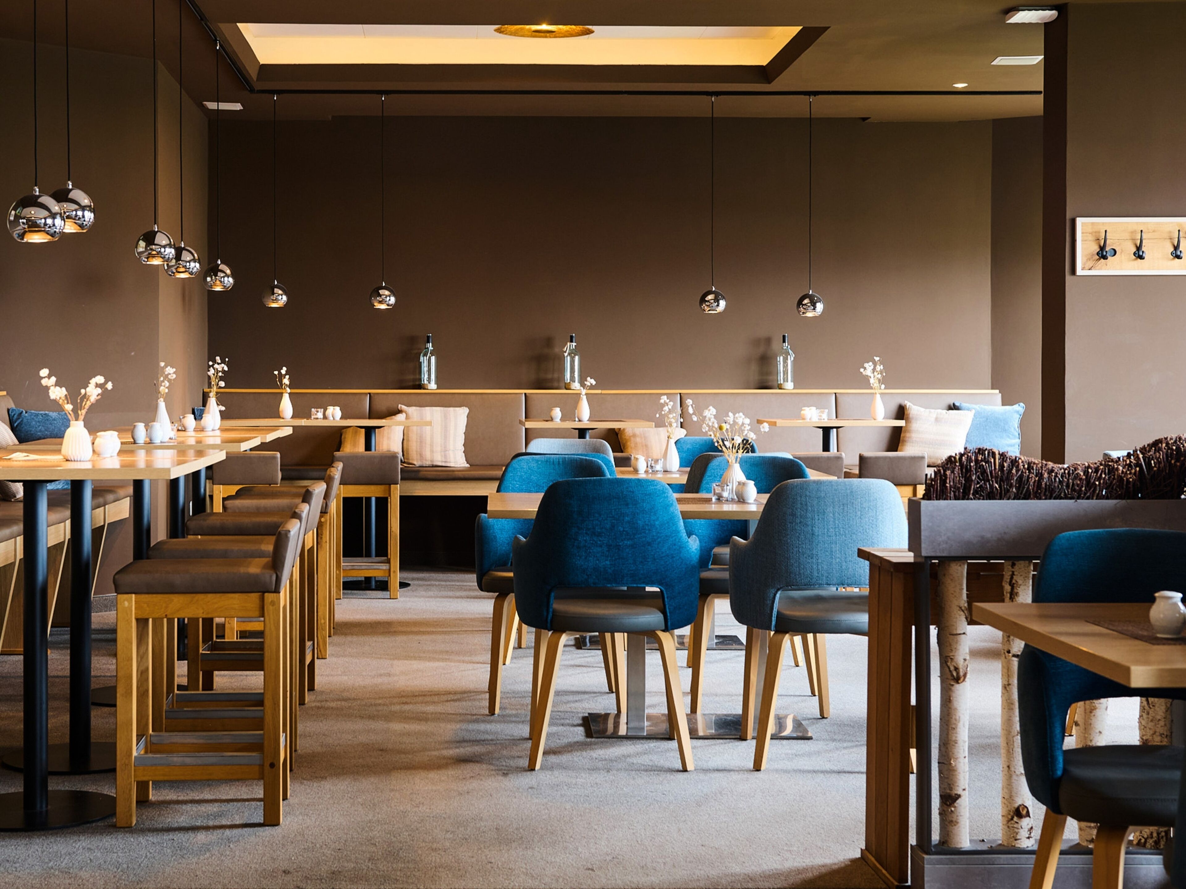 A modern restaurant interior with wooden tables, blue upholstered chairs, and hanging pendant lights.