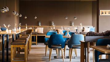 A modern restaurant interior with wooden tables, blue upholstered chairs, and hanging pendant lights.