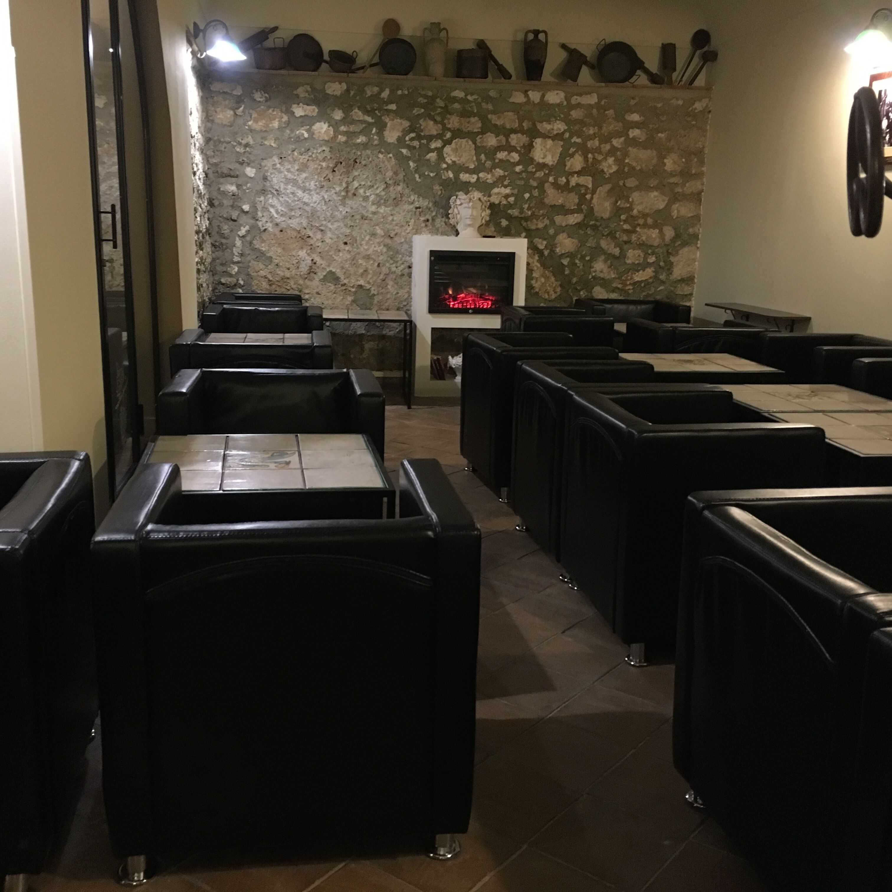 Un restaurante con cabinas de cuero negro dispuestas en filas, una pared de piedra con utensilios de cocina y una chimenea con un reloj digital.