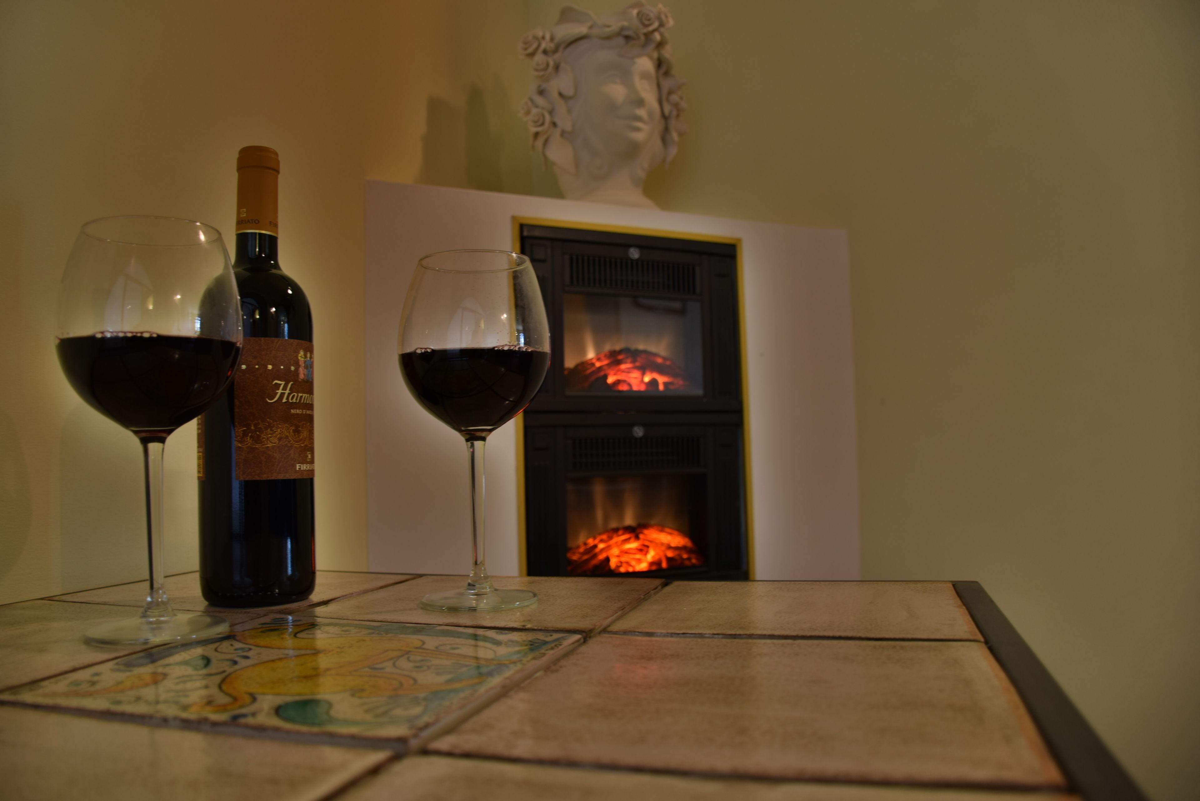 A table with two wine glasses and a bottle of wine, a fireplace, and a bust sculpture.