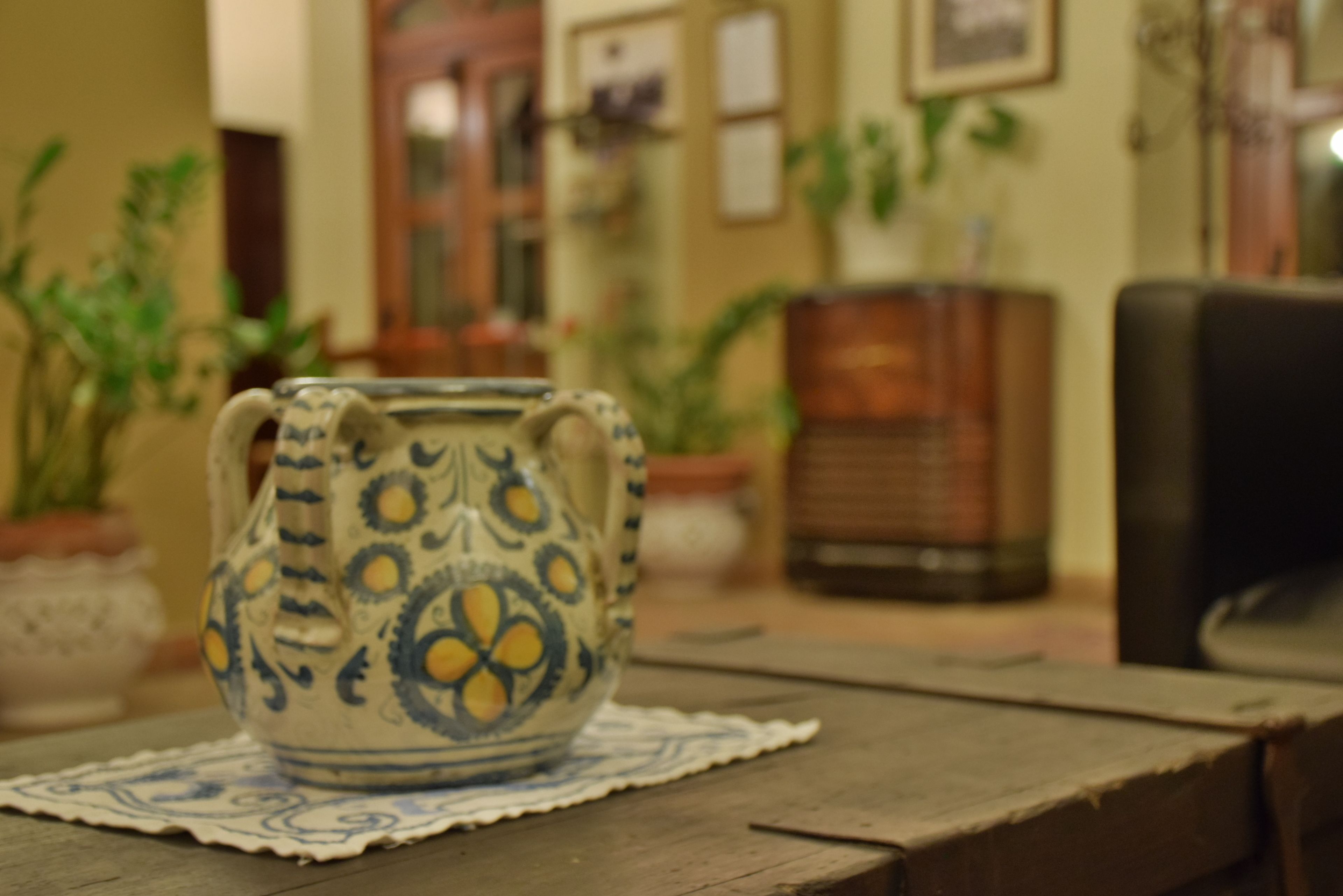A ceramic pitcher on a wooden table with a lace doily, surrounded by potted plants and furniture in a room with a traditional decor.