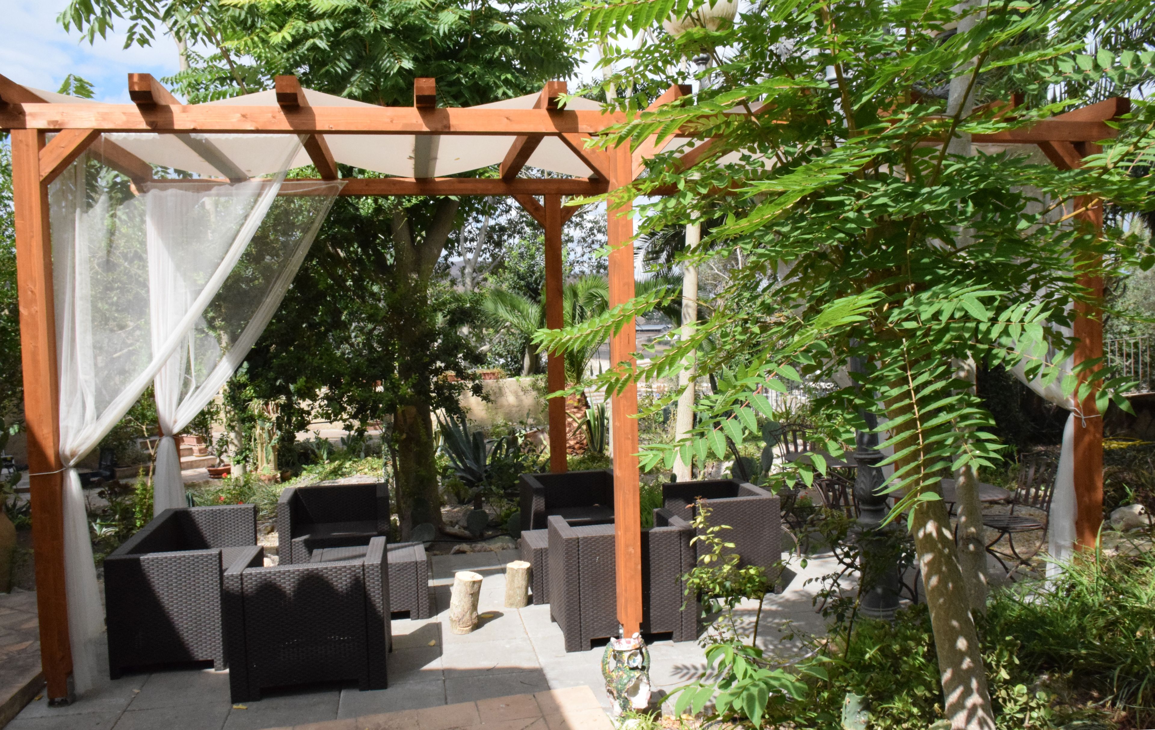 Un área de estar al aire libre con mobiliario de mimbre bajo una pérgola con toldo blanco.