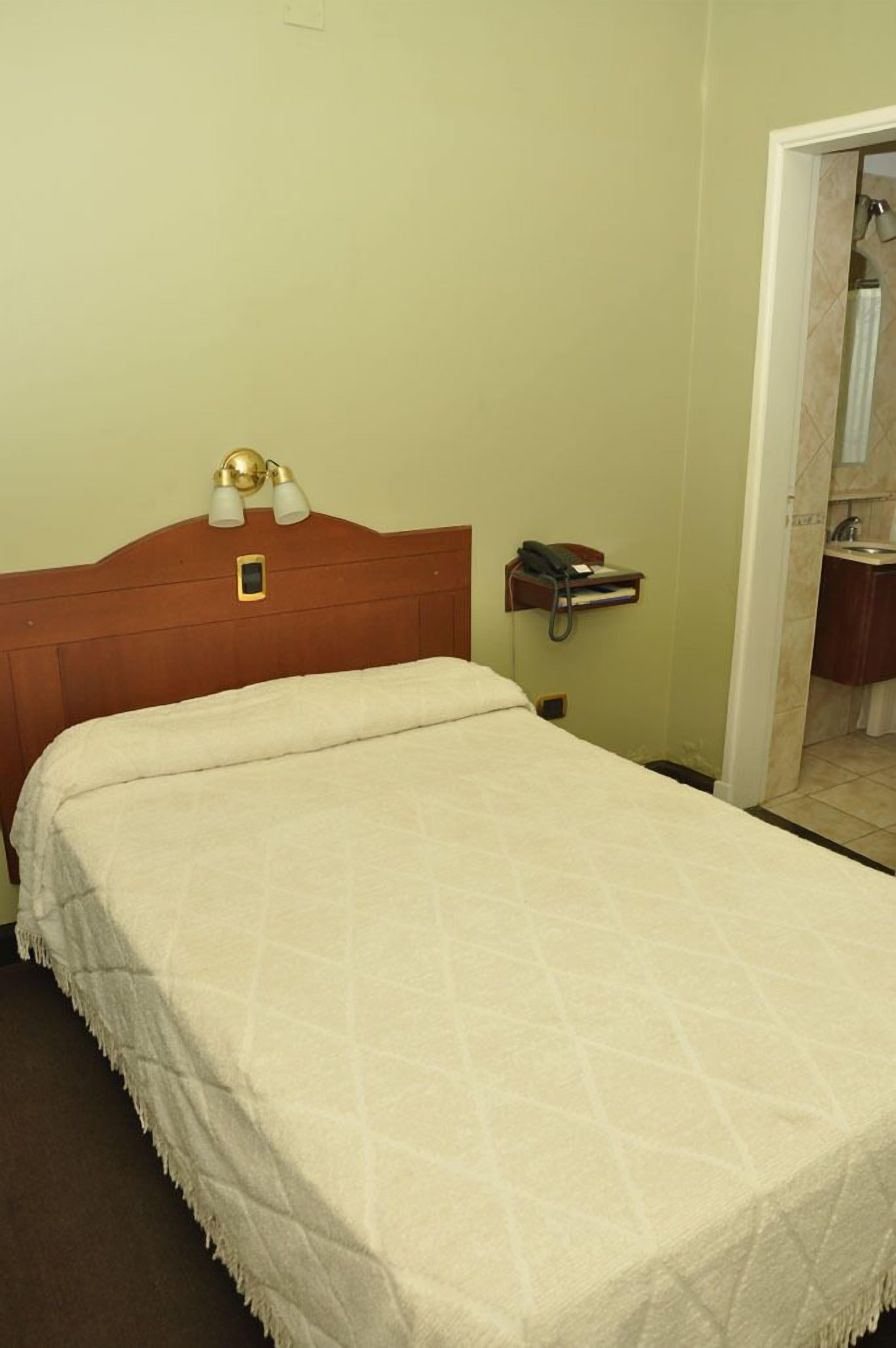 Habitación de hotel con una cama, una mesita de noche con lámpara, teléfono y un pequeño estante con un libro.