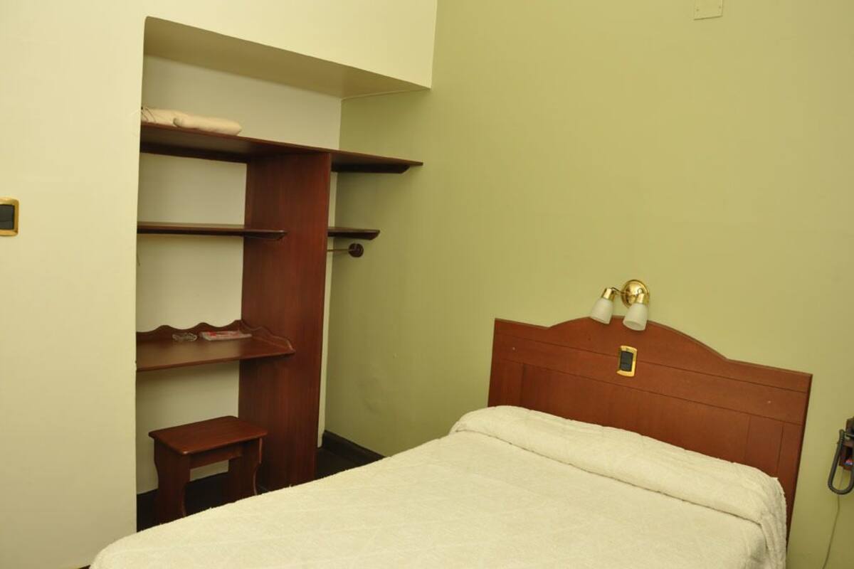 A bedroom with a bed, a wooden wardrobe, a small table, and a nightstand with a lamp.