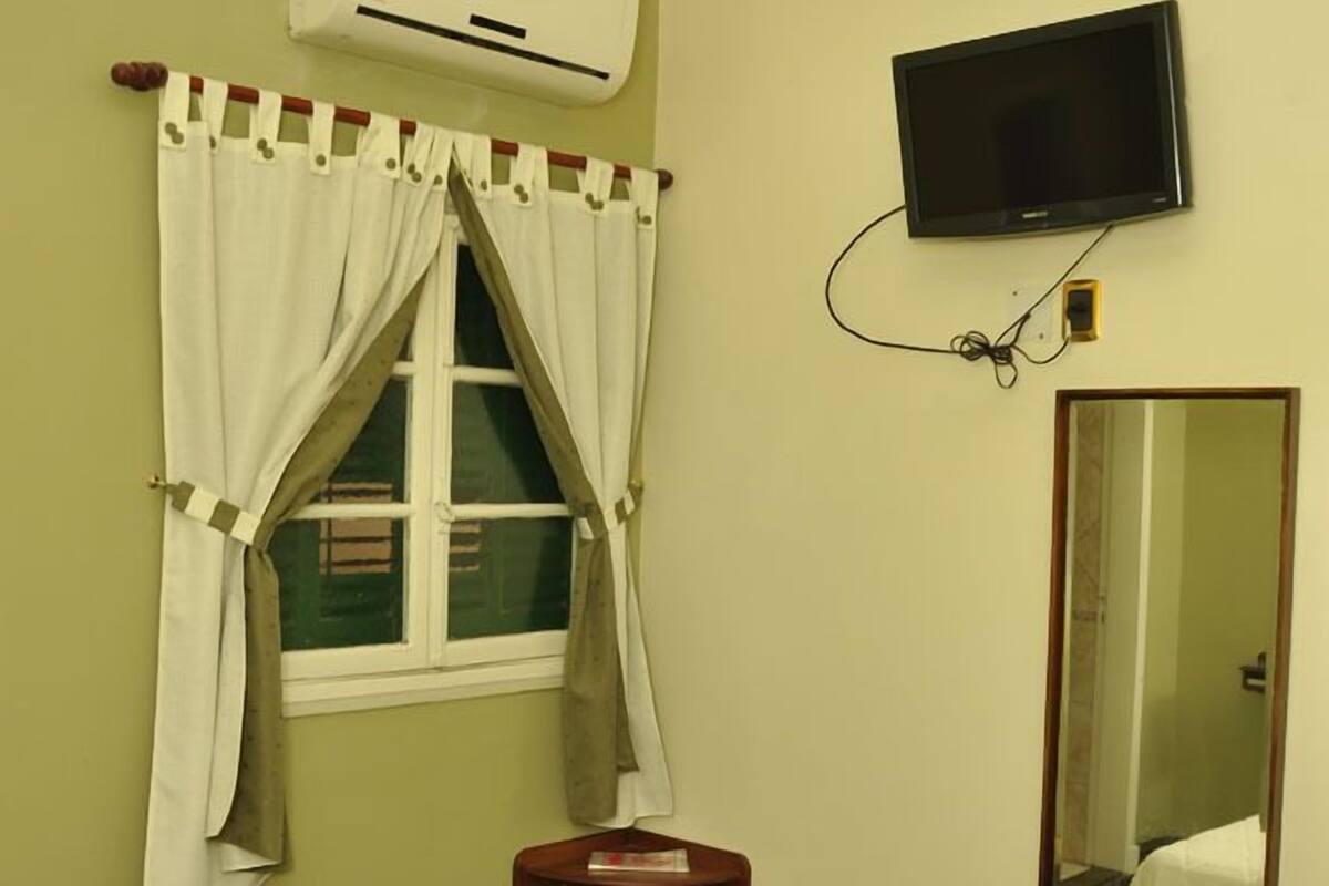 A room with a bed, an air conditioning unit, a television, a mirror, and a window with curtains.
