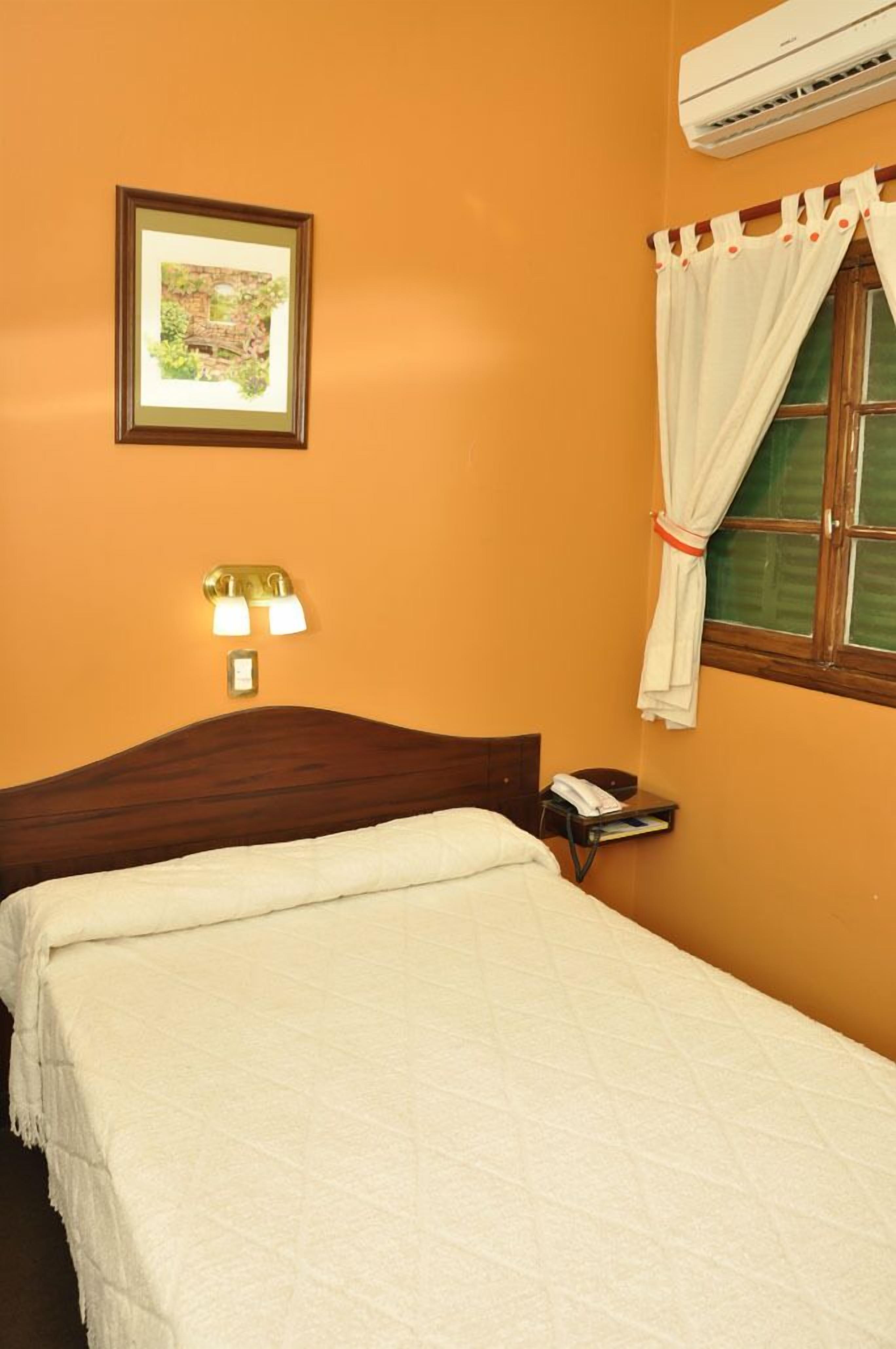 A hotel room with a bed, a nightstand, a framed picture on the wall, and an air conditioning unit.