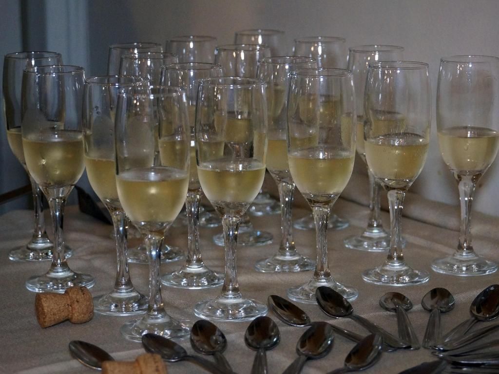 Vários copos de vinho cheios com uma bebida clara, dispostos sobre uma mesa com colheres e uma rolha.