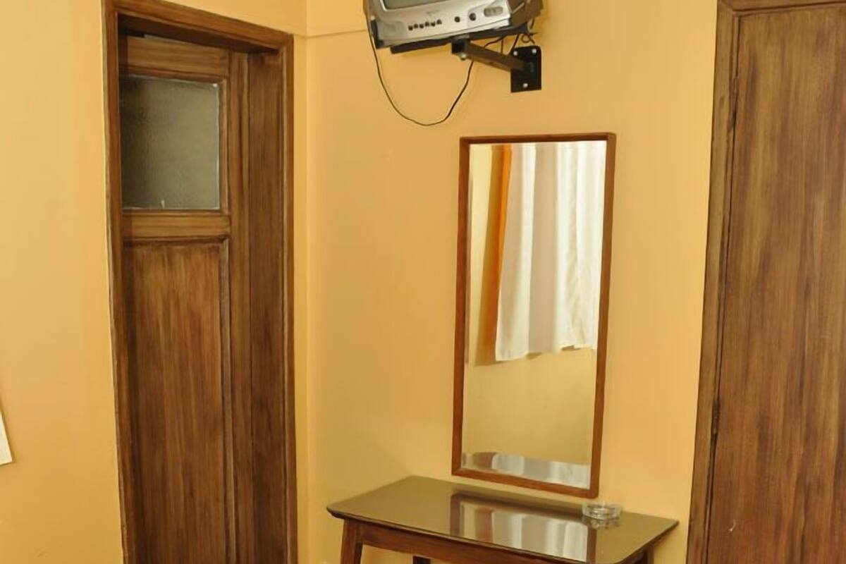 A hotel room with a bed, a wooden wardrobe, a small table, a mirror, and a television mounted on the wall.