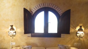 A room with two beds, a window with open shutters, and wall-mounted lamps.