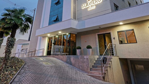 A modern hotel with a prominent 'Slaviero' sign, a staircase leading to the entrance, and a paved area with a palm tree.