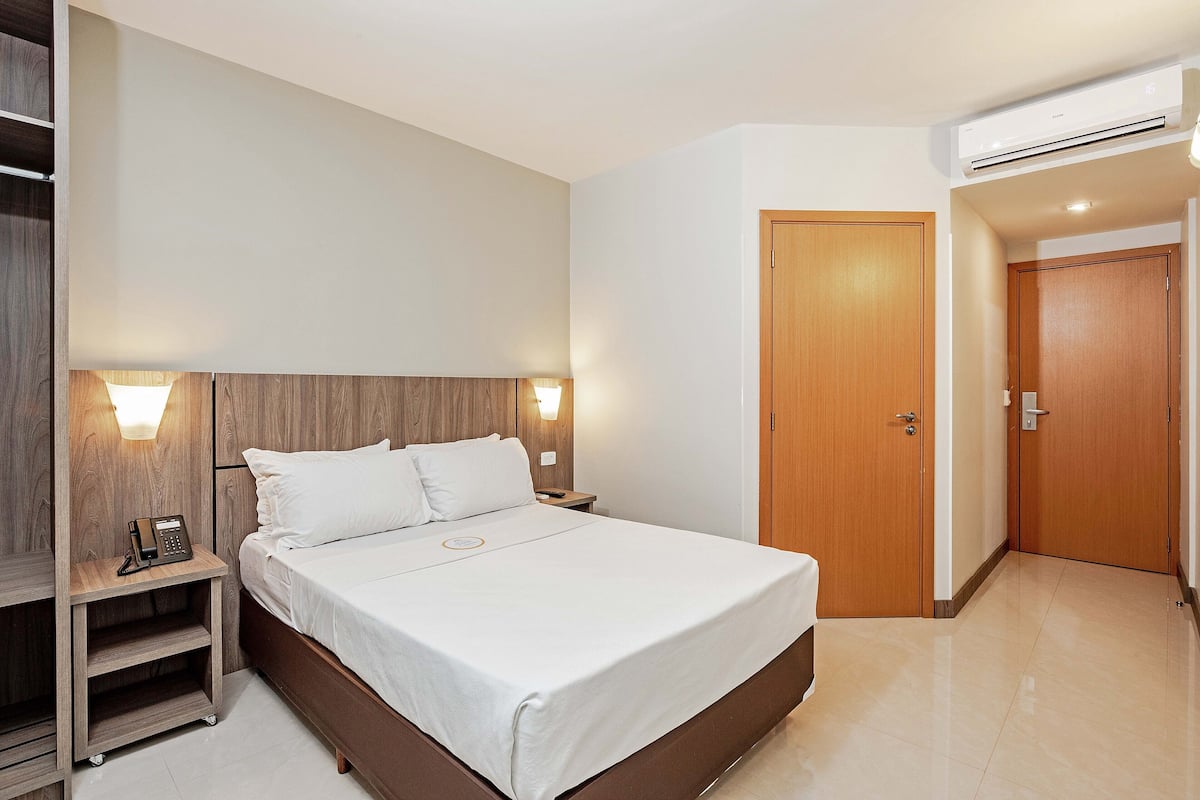 Hotellihuone, jossa on sänky, yöpöydät, puinen pääty, seinään kiinnitetty puhelin ja ovi.