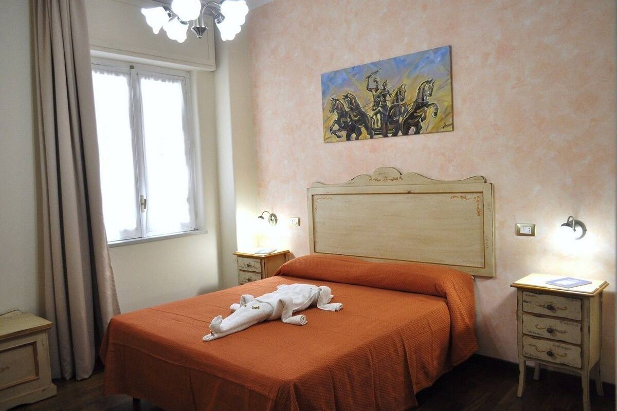 A bedroom with a bed, a nightstand, a lamp, a painting, and a window with curtains.