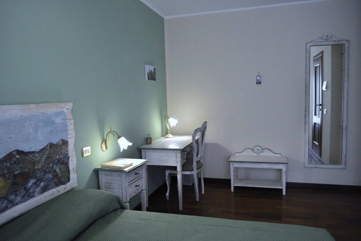 A bedroom with a bed, a desk with a chair, a small bedside table, a wall-mounted lamp, and a large mirror.