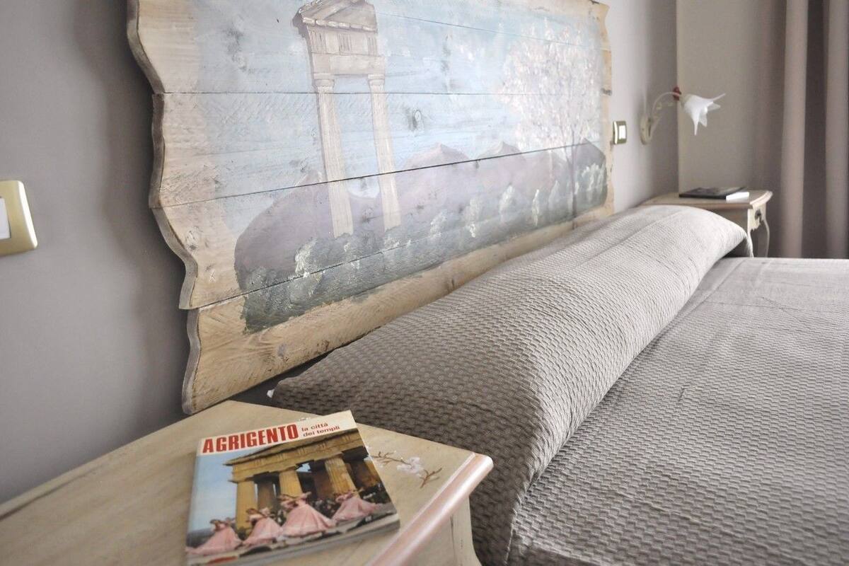 A bedroom with a large, rustic wall art piece depicting a temple and columns, a neatly made bed with a textured blanket, and a small bedside table with a book titled "AGRIGENTO."