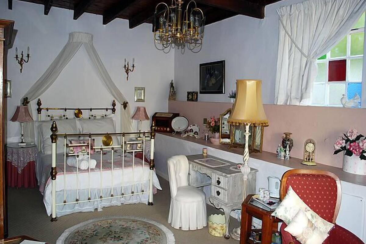 A bedroom with a four-poster bed, a bedside table, a chair, a desk, and a window with curtains.
