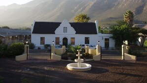 A white building with a black roof, a fountain in front, and mountains in the background. - Kingna Lodge (Montagu)