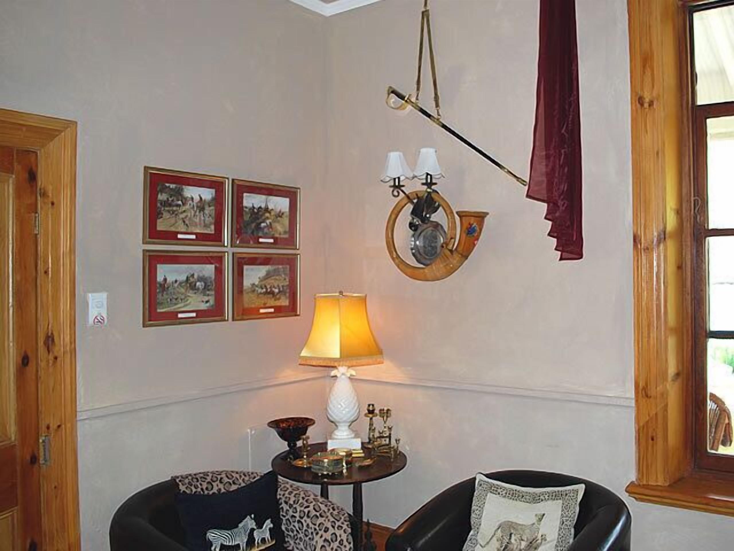 A cozy room with a wooden door, a table with a lamp, four framed pictures on the wall, and two chairs with patterned cushions.
