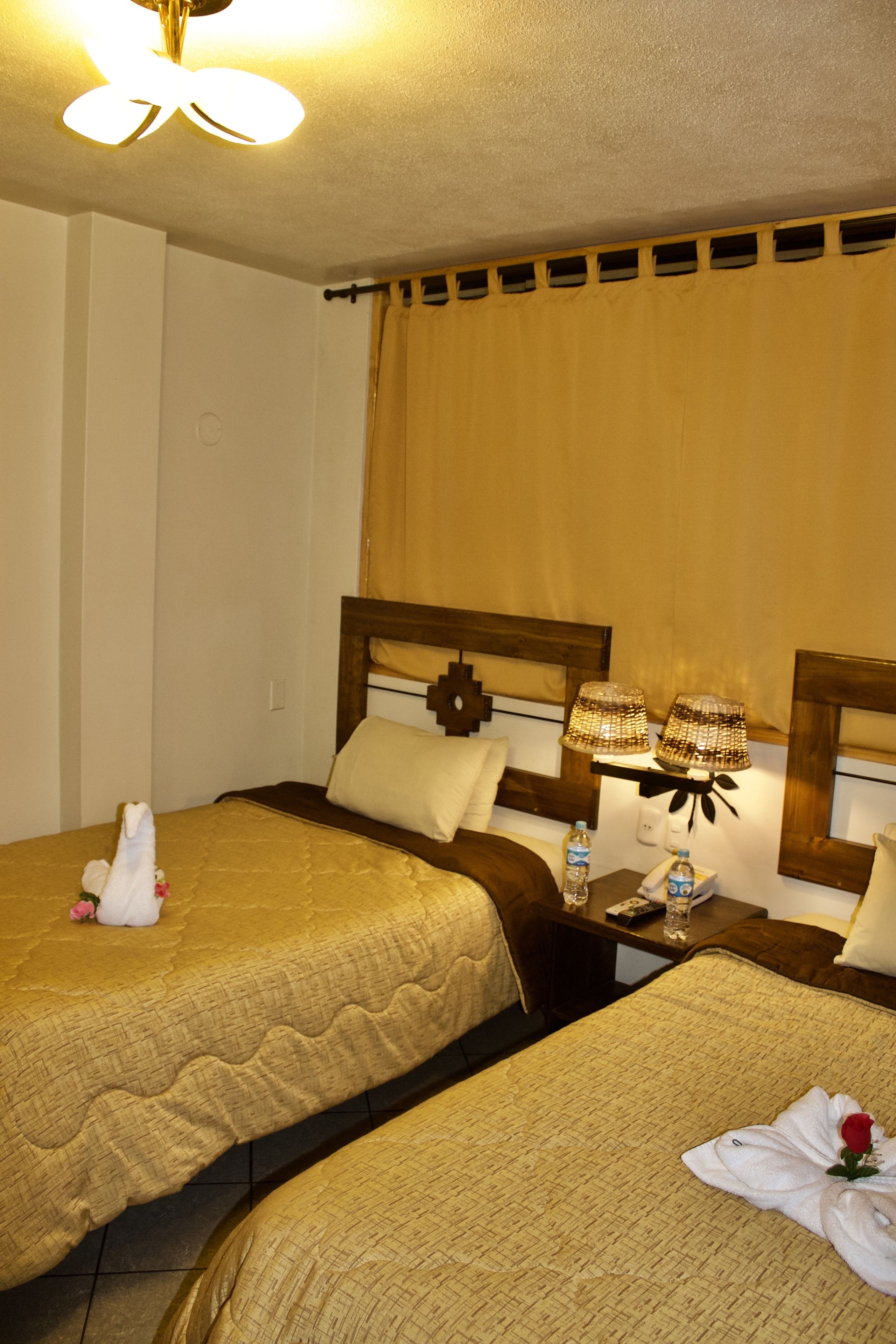 Habitación de hotel con dos camas, cada una con cabecero de madera y colcha acolchada color beige. Hay dos mesitas de noche, una con lámparas, una botella de agua y un control remoto.
