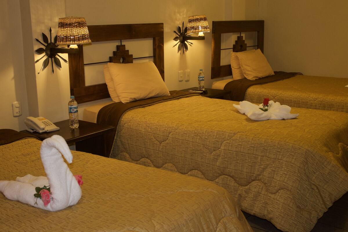 Habitación de hotel con dos camas, cada una con cabecero, mesita de noche y lámpara. Hay una botella de agua en la mesita y una toalla con forma de cisne y rosas sobre una de las camas.