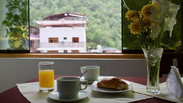 Une table de petit-déjeuner avec un croissant, deux tasses, un verre de jus d’orange et un vase rempli de fleurs, placé près d’une fenêtre donnant sur un immeuble.