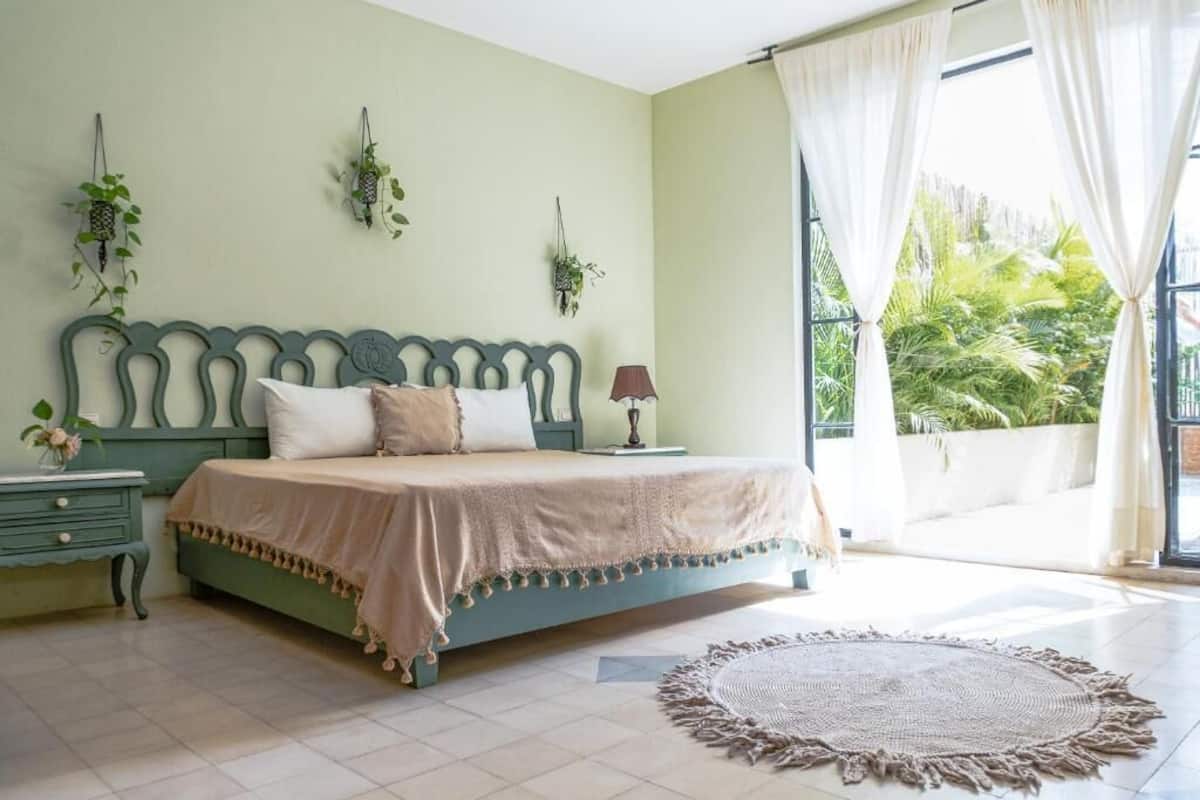 A bedroom with a bed, bedside table, lamp, and a view of greenery outside.