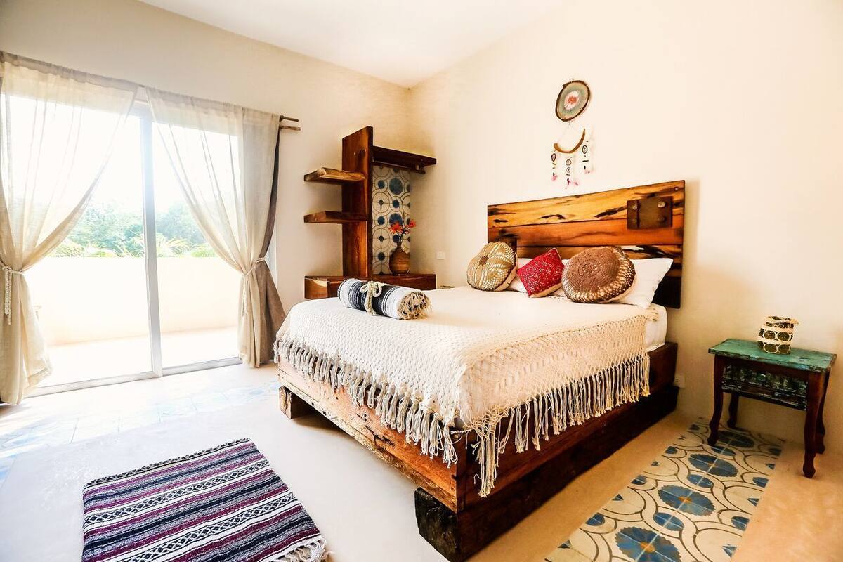 A bedroom with a large bed, a wooden headboard, a small table, and a view of the outdoors through a sliding door.