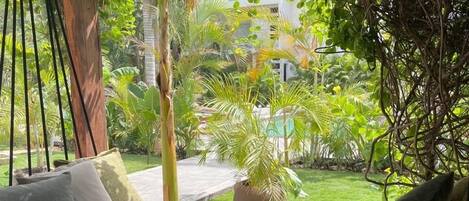 Una hamaca con almohadones, piso de piedra y vista a un jardín con diversas plantas y un edificio al fondo.