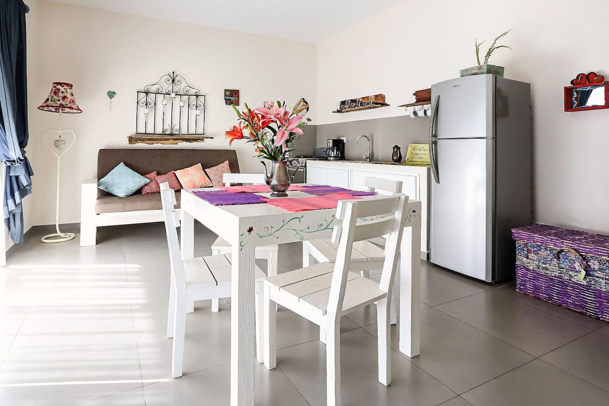 A compact living space with a dining table, white chairs, a sofa with cushions, a refrigerator, a small kitchen area, and a decorative plant.
