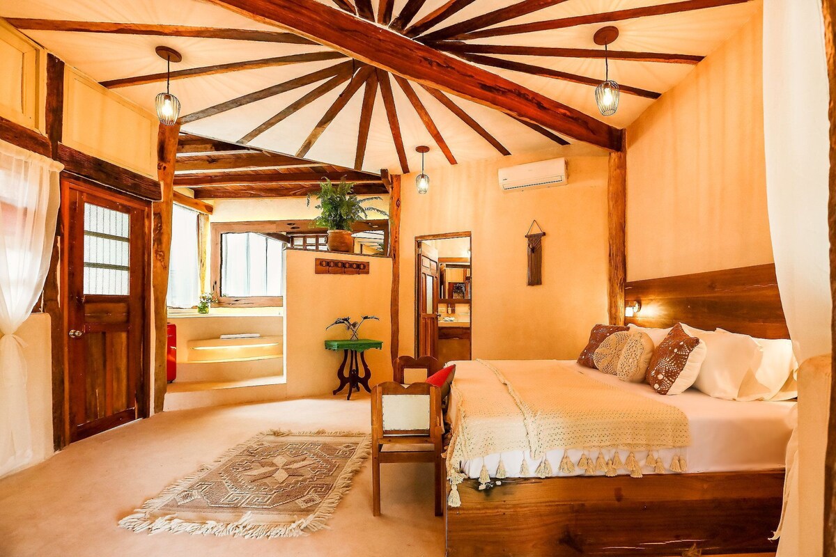 Une chambre avec un plafond en bois, un lit avec des oreillers, une chaise, une petite table et une plante.