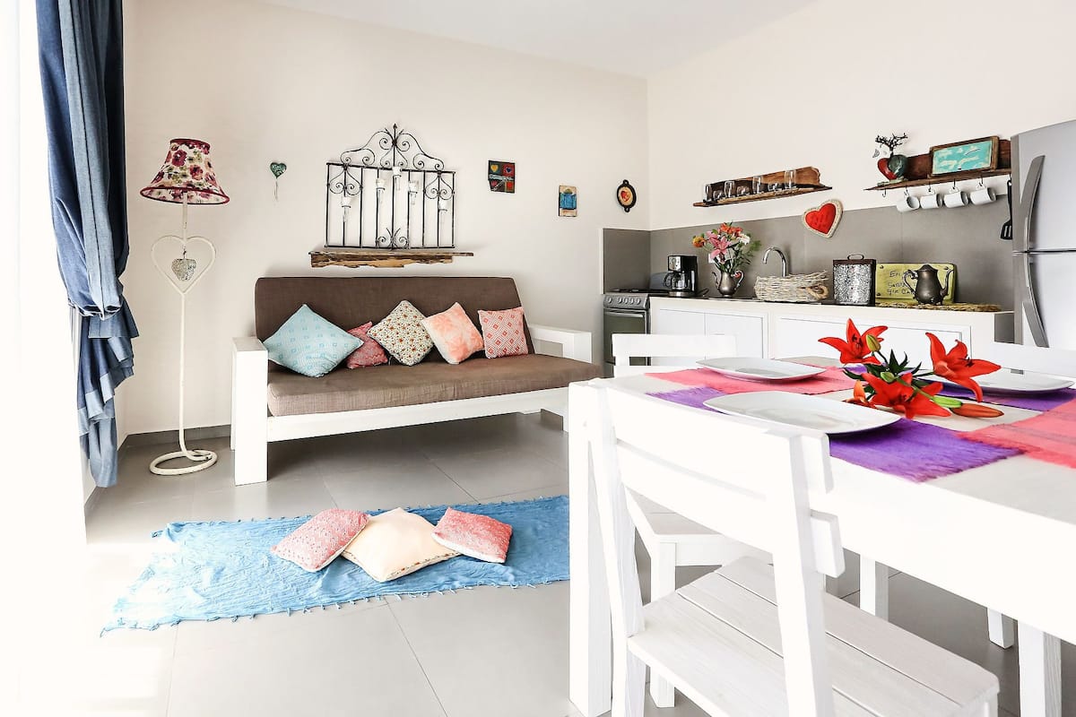 A modern kitchen with white cabinetry, a dining table with a colorful placemat, and a sofa with patterned cushions.
