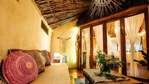 A cozy room with a woven ceiling, a sofa with a crocheted pillow, a wooden table with a vase of flowers, and a large window with sheer curtains. - Xscape Tulum (Tulum)