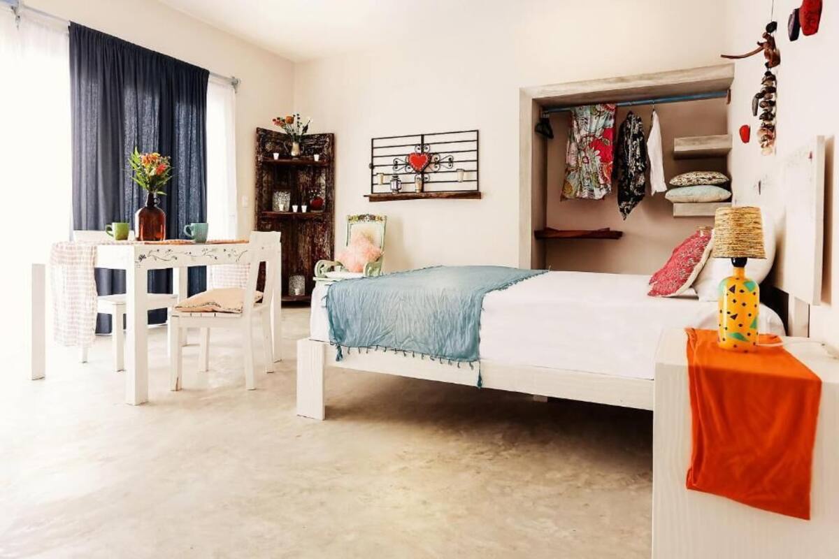 A bedroom with a bed, a desk, and a chair. There is a wardrobe with clothes and a mirror.