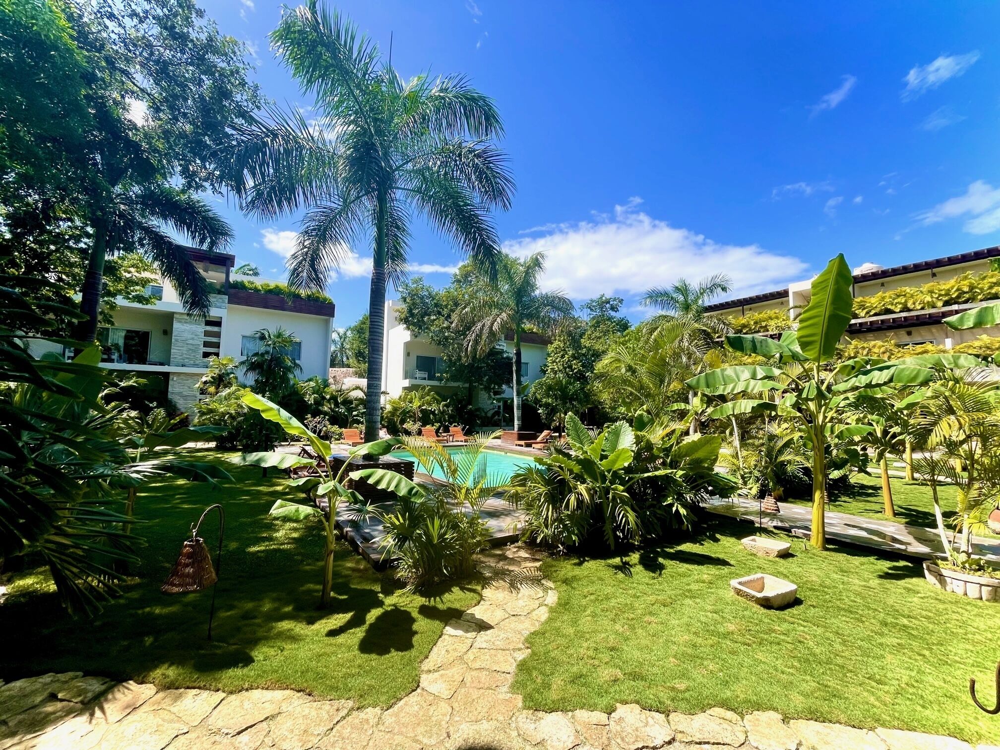 A well-maintained garden with a swimming pool, palm trees, and a stone pathway leading to a building with multiple balconies.