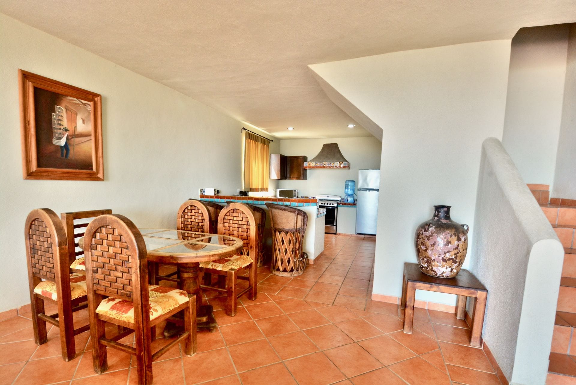 A dining area with a round table and six chairs, a kitchen in the background, and a staircase leading to another level.