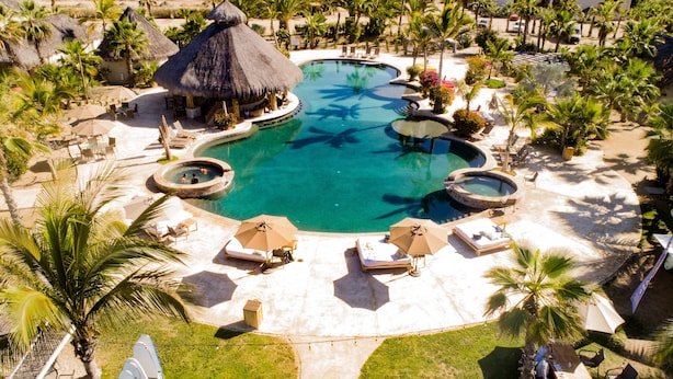 An aerial view of a resort with a large central pool, multiple lounge areas with umbrellas, and thatched-roof structures. - Cerritos Surf Town Beach Front Hotel and SPA (El Pescadero)