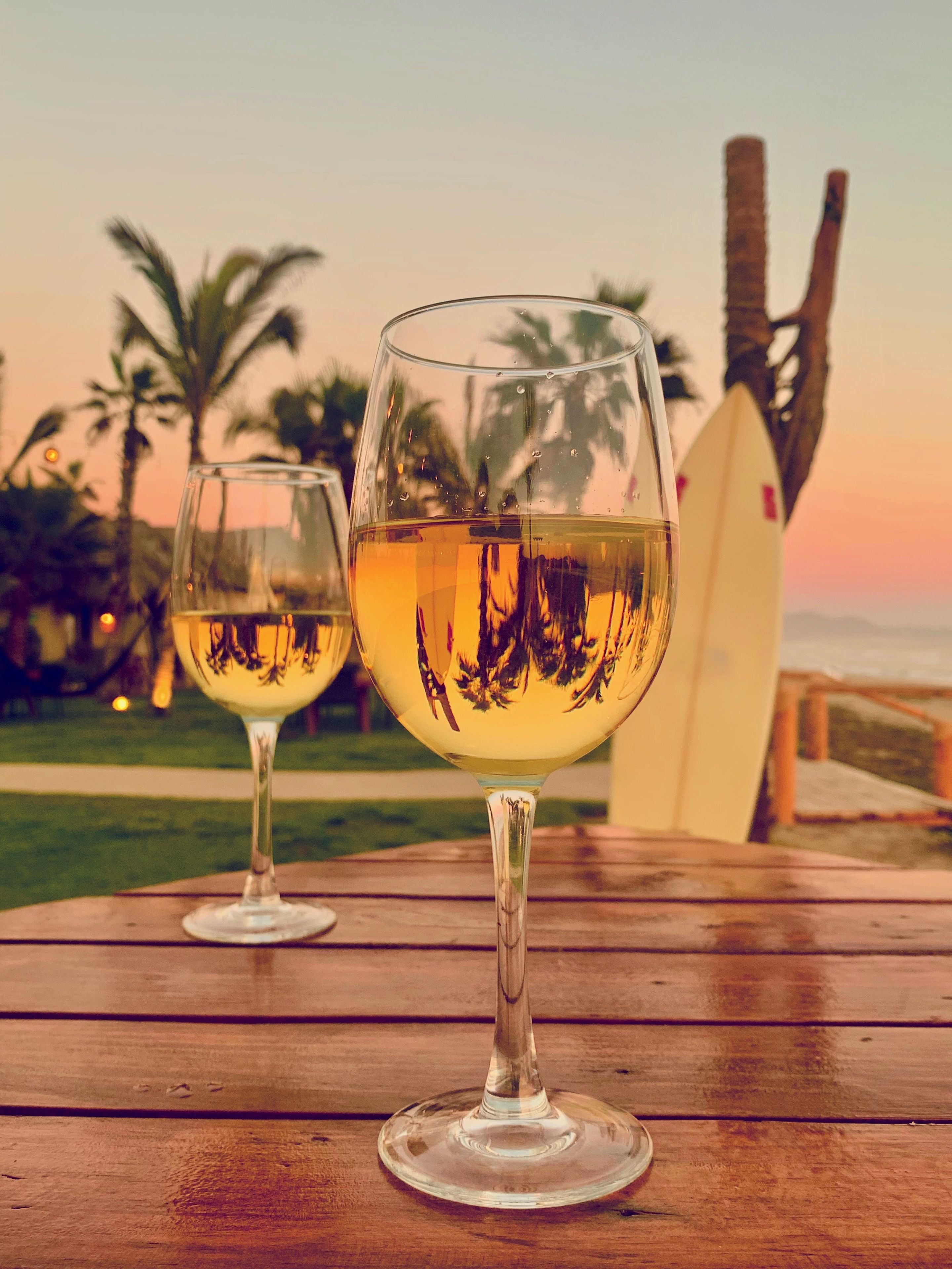 Two wine glasses on a wooden table with a surfboard and palm trees in the background.