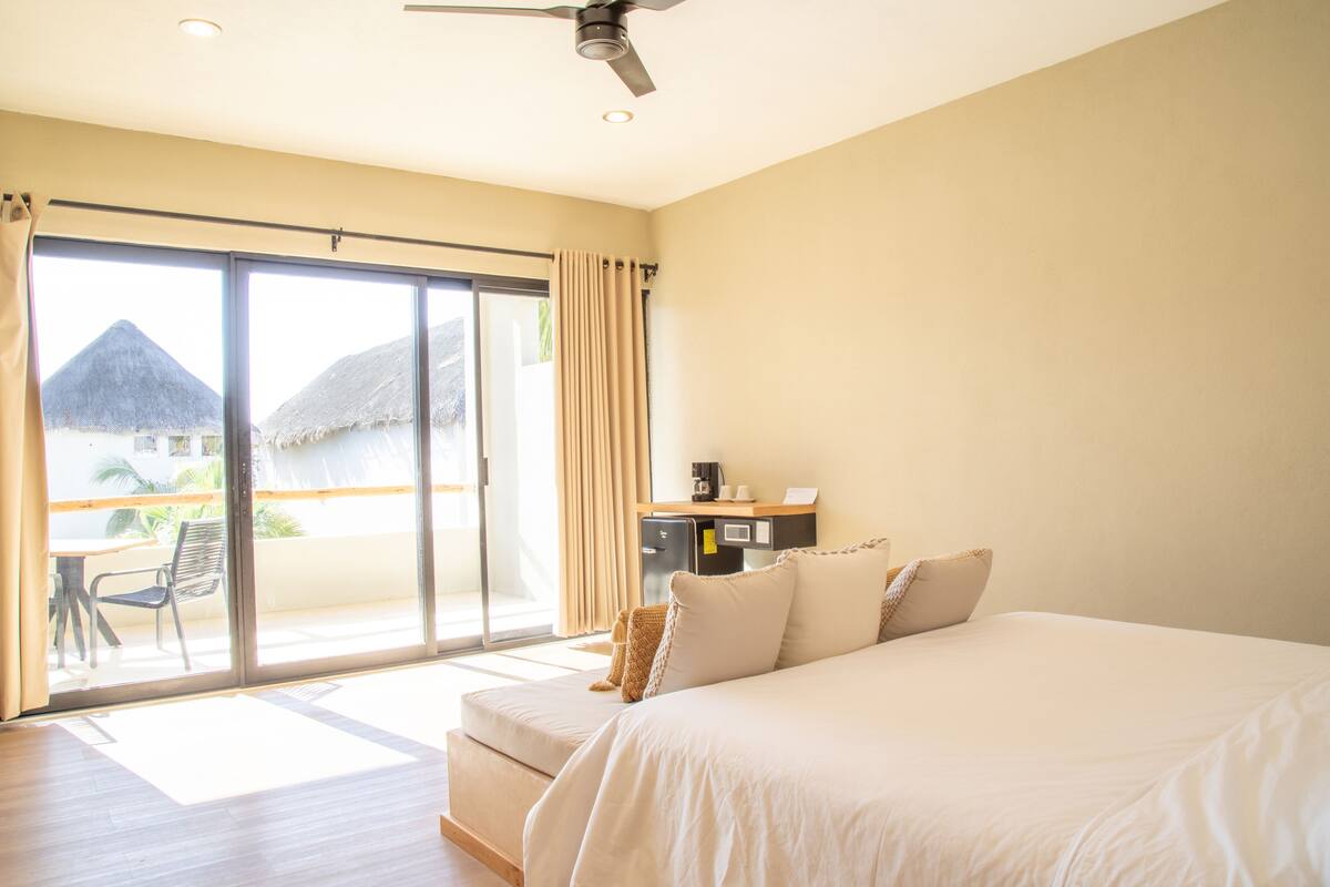 Une chambre d’hôtel avec un grand lit, un bureau avec un ordinateur, une chaise et une vue sur un bâtiment à toit de chaume, visible à travers une porte-fenêtre coulissante.