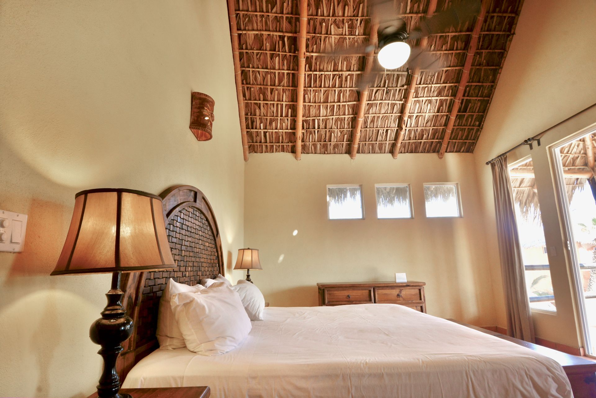 A bedroom with a bed, two bedside lamps, a wooden headboard, a ceiling fan, and a window with curtains.