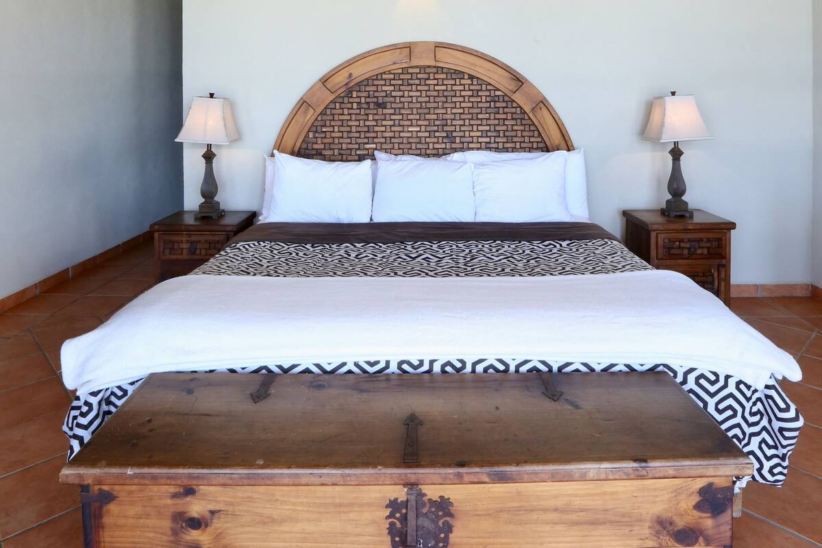 Une chambre à coucher avec une tête de lit en bois, un grand lit recouvert d’une couvre-lit à motifs, deux tables de chevet avec des lampes et un ventilateur de plafond.