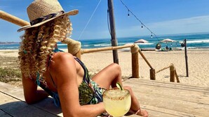 A person sitting on a wooden deck at a beach, holding a drink. - Cerritos Surf Town Beach Front Hotel and SPA (El Pescadero)