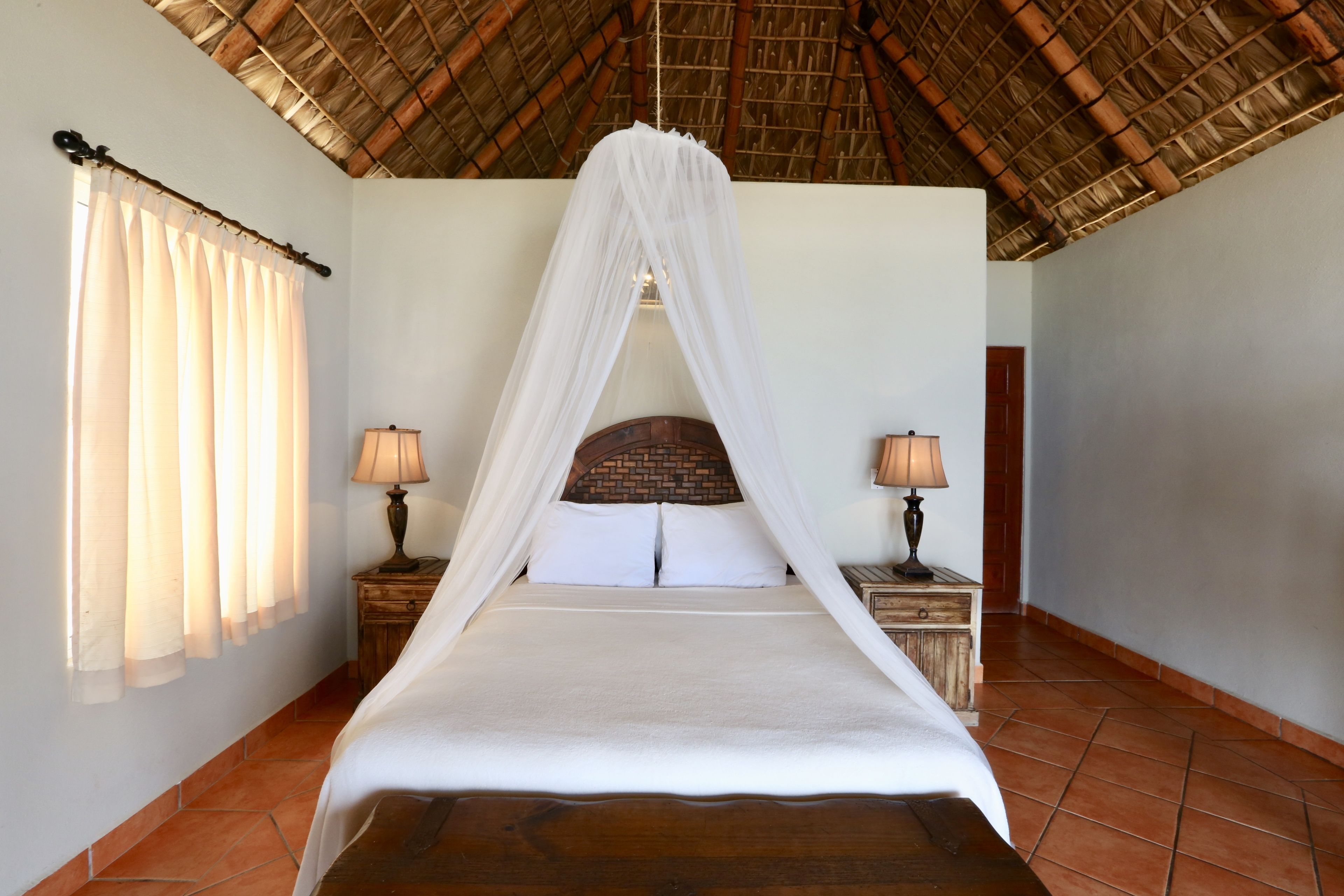 A bed with a canopy, two bedside tables with lamps, a window with curtains, and a tiled floor.