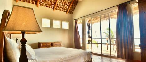 A bedroom with a bed, a nightstand, a lamp, a wooden dresser, a window with curtains, and a view of the outside.