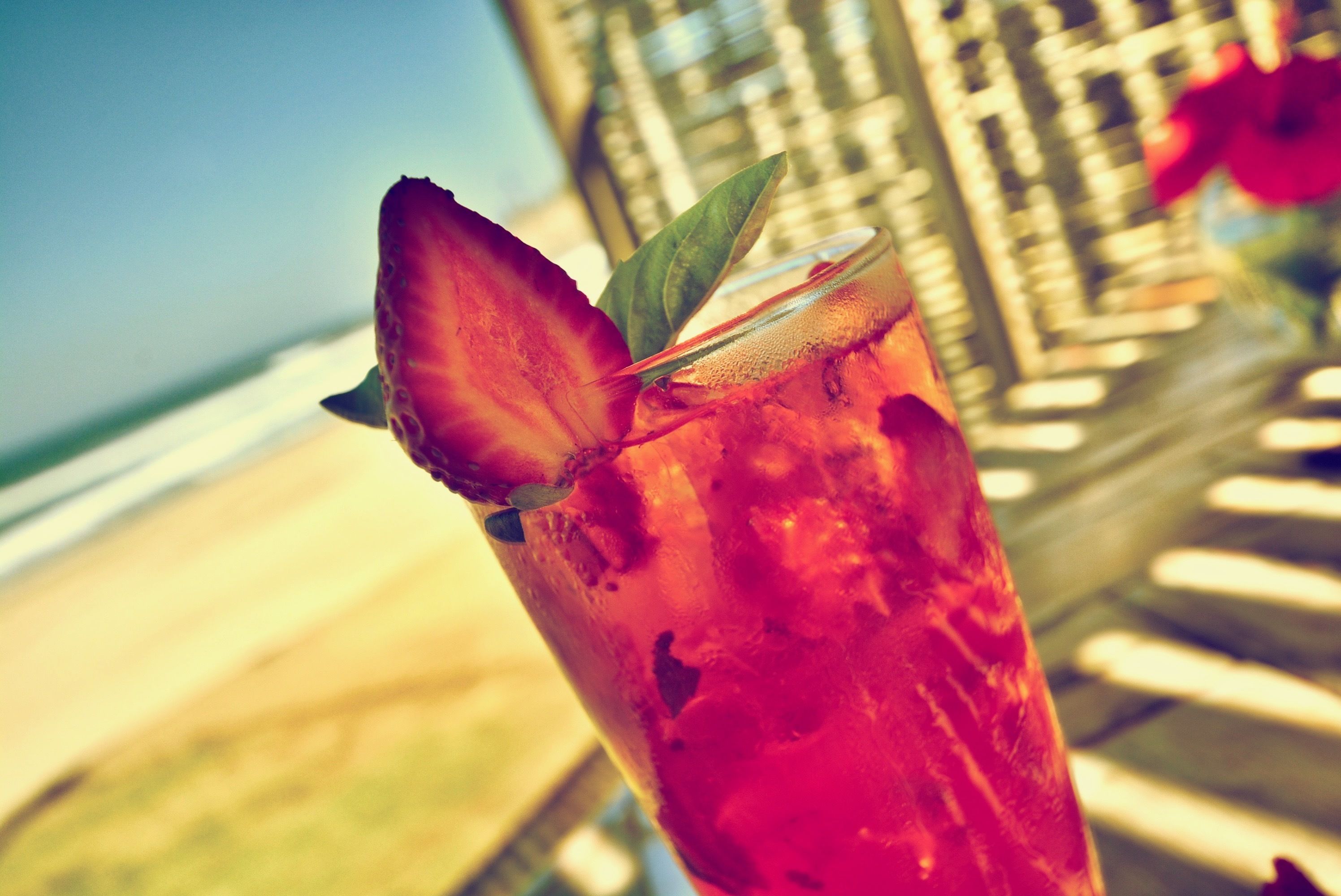 A cocktail with a strawberry garnish and a leaf, set against a beach and hotel background.