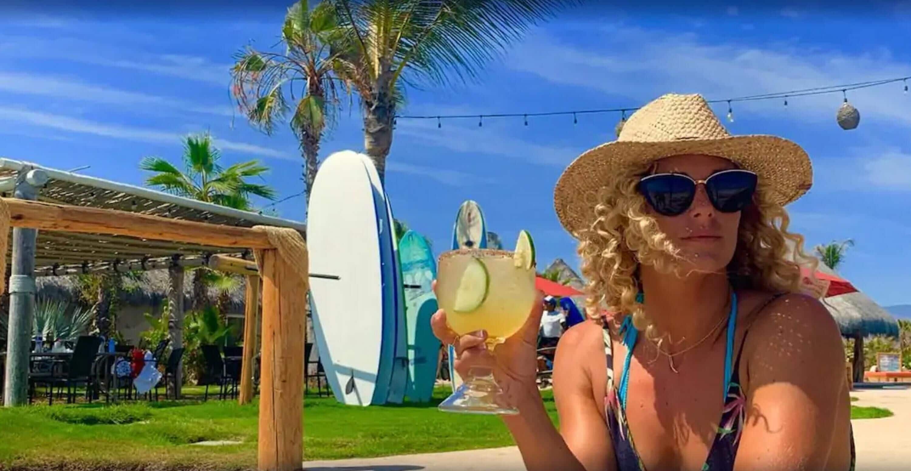A person holding a drink with a tropical background, including palm trees and a surfboard.