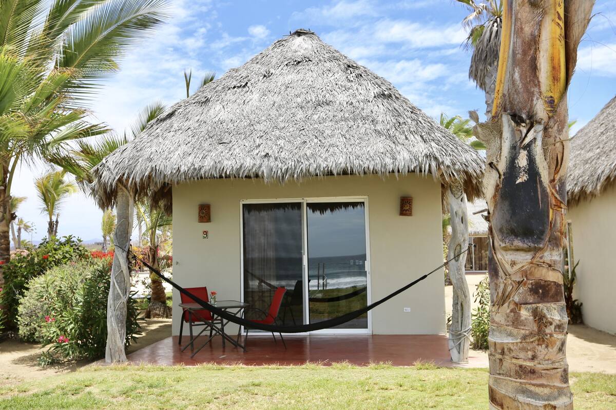Une structure à toit de chaume dotée d’une porte coulissante en verre, d’un hamac et d’une terrasse avec des chaises.