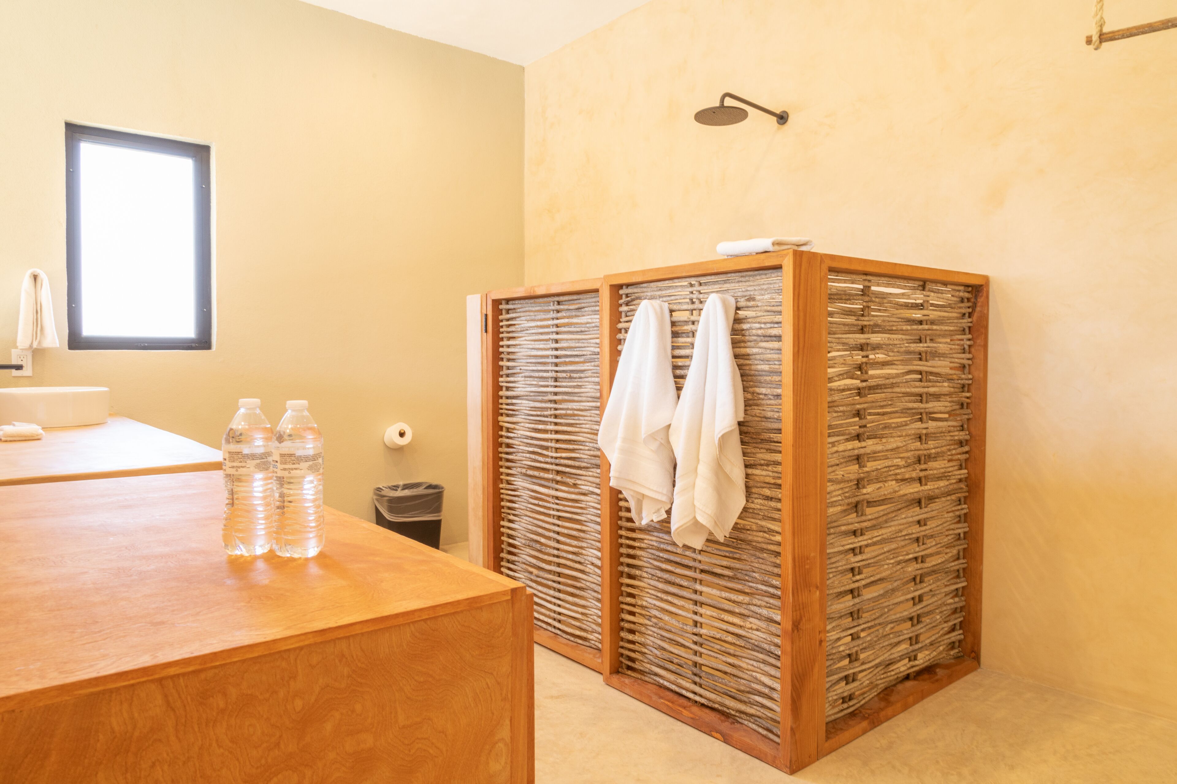 Ein Badezimmer mit einem Holzbadezimmermöbel, zwei Wasserflaschen, einem Handtuchhalter mit Handtüchern, einer Duschkabine und einem Fenster.